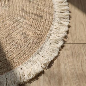 May include: A round, natural fiber rug with a woven texture and a cream-colored fringe border. The rug is a neutral beige color and appears to be made of jute or a similar material. The rug is on a light-colored wooden floor.