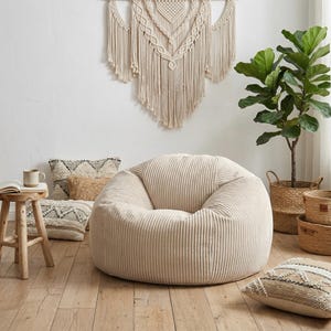 May include: A large, beige corduroy beanbag chair dominates a relaxed living space. Decorative cushions, a small wooden stool, and a macrame wall hanging contribute to the room's bohemian atmosphere. A potted plant and woven baskets complete the scene.