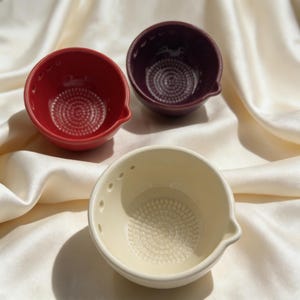 Peut inclure: Trois petits bols en céramique de couleurs rouge, violet et crème. Chaque bol a un intérieur texturé et un petit bec verseur. Les bols sont disposés sur une surface blanche et soyeuse. Ces bols sont parfaits pour la cuisine.