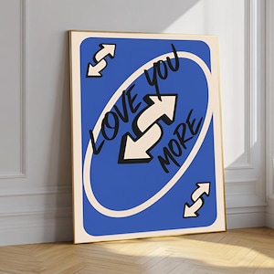 May include: A framed art print featuring a blue card design with the words "LOVE YOU MORE" in black and white. The artwork includes white arrows and a cream-colored border, set against a gold frame. The print is displayed on a wooden floor.