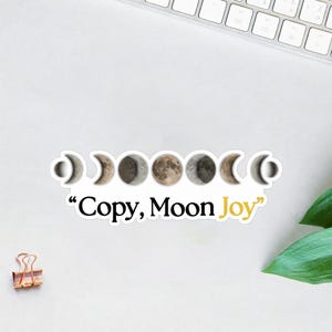 May include: A white sticker featuring the phases of the moon in a row. The sticker has the text "Copy, Moon Joy" in black and yellow. The sticker is on a white surface with a keyboard and green leaves in the background.