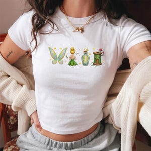 Könnte beinhalten: Ein weißes Kurzarm-T-Shirt mit einem Grafikdruck von Feenflügeln, einer kleinen Figur mit blondem Dutt, einer leuchtenden Zaubertrankflasche und einem grünen, spulenförmigen Haus mit einem roten Pilz darauf, akzentuiert durch kleine gelbe Sterne.