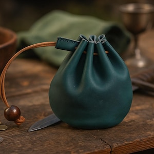 Può includere: Un sacchetto con coulisse in pelle verde acqua con cordoncino in pelle marrone e perla di legno. Il sacchetto è rotondo e poggia su una superficie di legno, con un coltello e monete nelle vicinanze.