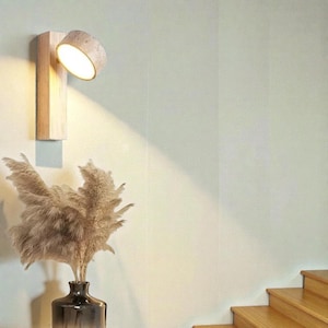 Op de afbeelding: Een houten wandlamp met een cilindrische lamp. Het licht is naar beneden gericht en geeft een warme gloed. De lamp is gemonteerd op een lichtgekleurde houten basis. Een vaas met gedroogd pampasgras staat eronder.