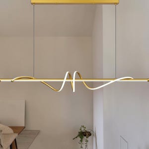 Può includere: Una moderna lampada a sospensione lineare color oro con luce LED bianca. L'apparecchio ha un design curvo con un anello centrale ed è sospeso al soffitto tramite sottili fili. La luce è accesa.