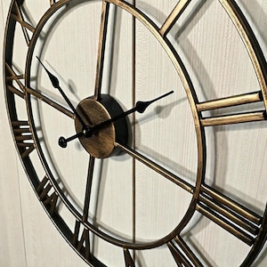 Può includere: Un grande orologio da parete rotondo con finitura oro metallizzato e lancette nere. Il quadrante dell'orologio presenta numeri romani e un design circolare. Le lancette dell'orologio indicano circa le 10:10. L'orologio è un oggetto decorativo.