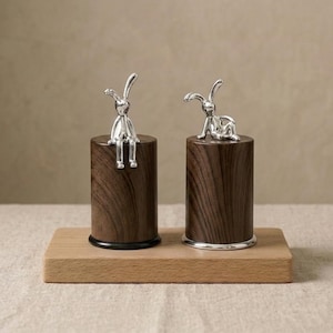 Puede incluir: Dos saleros y pimenteros decorativos. Cada uno presenta una figura de conejo plateado sobre una base cilíndrica de madera marrón oscuro. Las bases se asientan sobre una bandeja rectangular de madera. Un conejo está sentado, el otro está agachado.