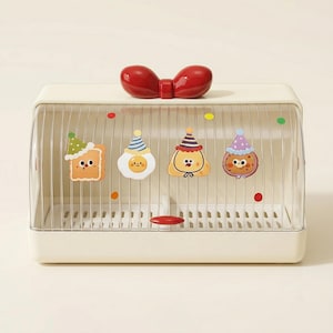 Puede incluir: Una panera rectangular de color crema con una parte delantera transparente y arqueada. Cuenta con un asa en forma de lazo rojo y cuatro pegatinas de comida de dibujos animados con sombreros de fiesta. El interior es de rejilla metálica.