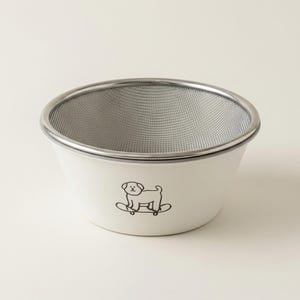 May include: A white ceramic strainer with a stainless steel mesh interior and rim. A black cartoon dog on a skateboard is printed on the side. The strainer is designed for kitchen use.