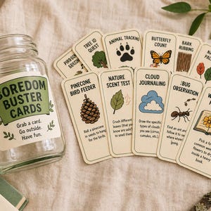 May include: A glass jar with a label reading "Boredom Buster Cards" and a collection of illustrated activity cards. The cards depict nature-based activities, including "Animal Tracking" and "Wild Flower Pressing". The jar is next to a stack of cards.