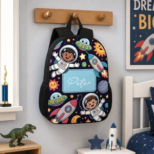 Może przedstawiać: Czarny plecak z motywem kosmicznym przedstawiającym astronautów, rakiety, latające spodki, planety i gwiazdy. Na środku znajduje się jasnoniebieska prostokątna naszywka z imieniem Peter napisanym białymi literami w stylu pisma odręcznego.
