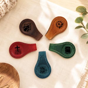 May include: Five teardrop-shaped leather magnetic bookmarks in dark brown, tan, red, green, and blue. Each bookmark features a unique black laser-engraved religious icon, including a church, a cross with a wreath, a cross with flowers, and a Bible.