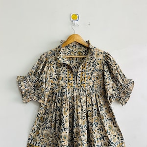 Könnte beinhalten: Eine kurzärmelige Bluse mit cremefarbener Basis und einem floralen Blockdruckmuster in Dunkelgrün und Senfgelb. Das Kleidungsstück hat einen Rüschenkragen, einen geschlitzten Ausschnitt und gerafften Stoff an der Taille für eine lockere Silhouette.