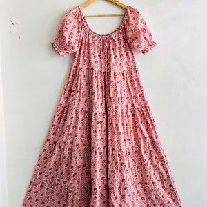 May include: A tiered midi dress in a light pink hue featuring a repeating dark pink floral block print pattern. The dress has short puff sleeves with elasticated cuffs, a wide scoop neckline, and a central drawstring tie at the chest.