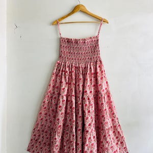 May include: A pink midi dress featuring a repeating floral pattern in a darker shade of pink. The garment has a square neckline, thin spaghetti straps, and a smocked bodice that transitions into a tiered, gathered skirt with a flared silhouette.
