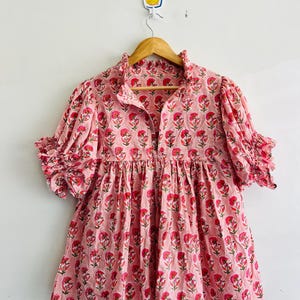 Puede incluir: Una blusa rosa claro con un patrón floral repetitivo en rosa oscuro y verde. La prenda tiene un cuello con volantes, cierre frontal de botones y mangas cortas abullonadas con puños con volantes. La silueta es holgada y fruncida en la cintura.