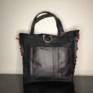 Pode incluir: Bolsa tote de couro preta com alça de metal prateado e detalhes em fita vermelha. A bolsa tem formato retangular com costuras verticais e uma parte superior aberta grande. As alças são resistentes e a bolsa parece espaçosa.