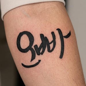 May include: A black tattoo on the arm featuring Korean characters. The tattoo is in a bold, handwritten style, with the characters prominently displayed against the skin. The arm is partially visible, with a neutral background.