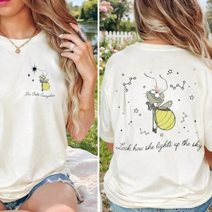 May include: Cream-colored t-shirt featuring a cartoon firefly with a yellow glowing abdomen. The front of the shirt has the text "Ma Belle Evangeline" and a star graphic. The back of the shirt has the text "Look how she lights up the sky".