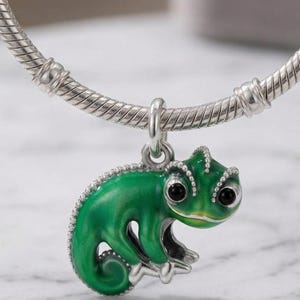 Puede incluir: Una pulsera de plata con un charm de camaleón verde. El camaleón tiene ojos negros y un acabado de esmalte verde detallado. La pulsera es de plata.