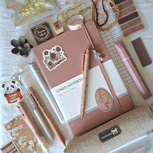 K&ouml;nnte beinhalten: Ein Set aus Schreibwaren, bestehend aus einem altrosa linierten Notizbuch, einem glitzernden ros&eacute;goldenen Stift, einem beigen Federm&auml;ppchen mit handgefertigtem Etikett, verschiedenen Blumen- und Panda-Aufklebern, einem Lesetagebuch, Washi-Tape und einem Lesezeichen.