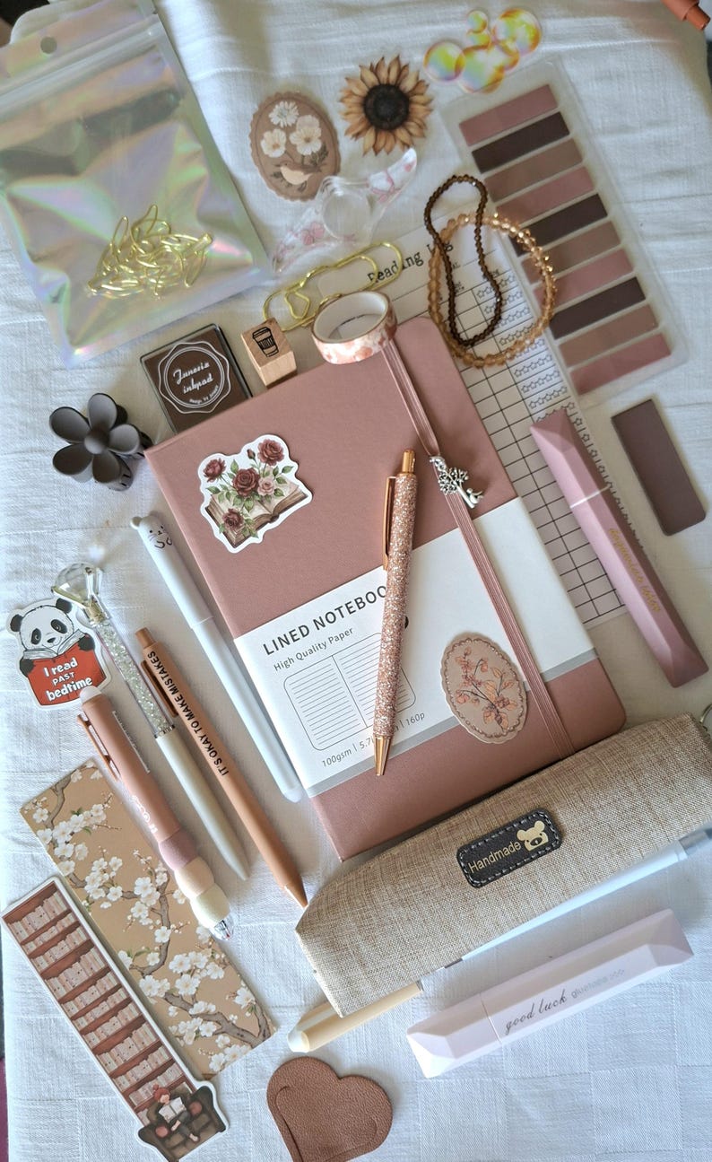 K&ouml;nnte beinhalten: Ein Set aus Schreibwaren, bestehend aus einem altrosa linierten Notizbuch, einem glitzernden ros&eacute;goldenen Stift, einem beigen Federm&auml;ppchen mit handgefertigtem Etikett, verschiedenen Blumen- und Panda-Aufklebern, einem Lesetagebuch, Washi-Tape und einem Lesezeichen.