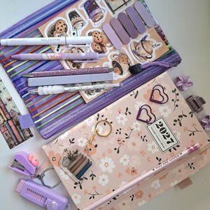 Könnte beinhalten: Ein violettes Netz-Etui mit Stiften, Aufklebern und Haftnotizen, neben einem floralen Planer für 2027, herzförmigen Büroklammern, einem Schlüsselanhänger, einem kleinen Hefter, einem Cuttermesser und einem Stift mit der Aufschrift YOU ARE LOVED.