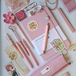 K&ouml;nnte beinhalten: Eine Sammlung rosa Schreibwaren, darunter ein Notizbuch mit Punkten, mehrere Stifte, ein Federm&auml;ppchen, eine Leseliste, Aufkleber, B&uuml;roklammern, ein blumenf&ouml;rmiger Clip, ein Perlenarmband und ein Lesezeichen mit einer Illustration einer lesenden Person.