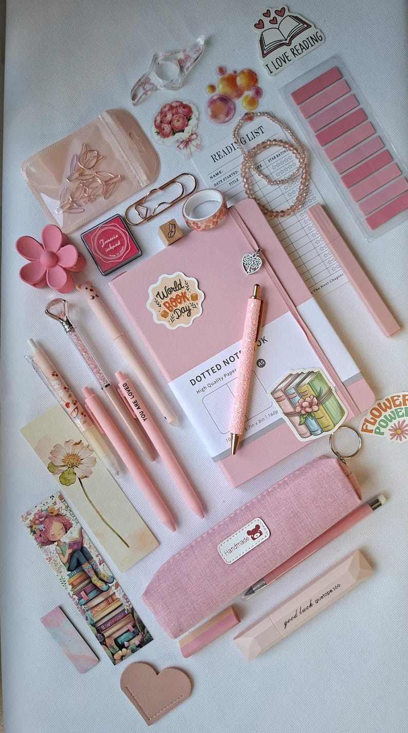 K&ouml;nnte beinhalten: Eine Sammlung rosa Schreibwaren, darunter ein Notizbuch mit Punkten, mehrere Stifte, ein Federm&auml;ppchen, eine Leseliste, Aufkleber, B&uuml;roklammern, ein blumenf&ouml;rmiger Clip, ein Perlenarmband und ein Lesezeichen mit einer Illustration einer lesenden Person.