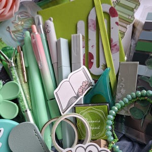 May include: A collection of stationery items in shades of green and pink, including a lime green notebook with an elastic band, various pens, a small ink pad, decorative washi tape, a beaded bracelet, stickers of books, and artificial pink roses.