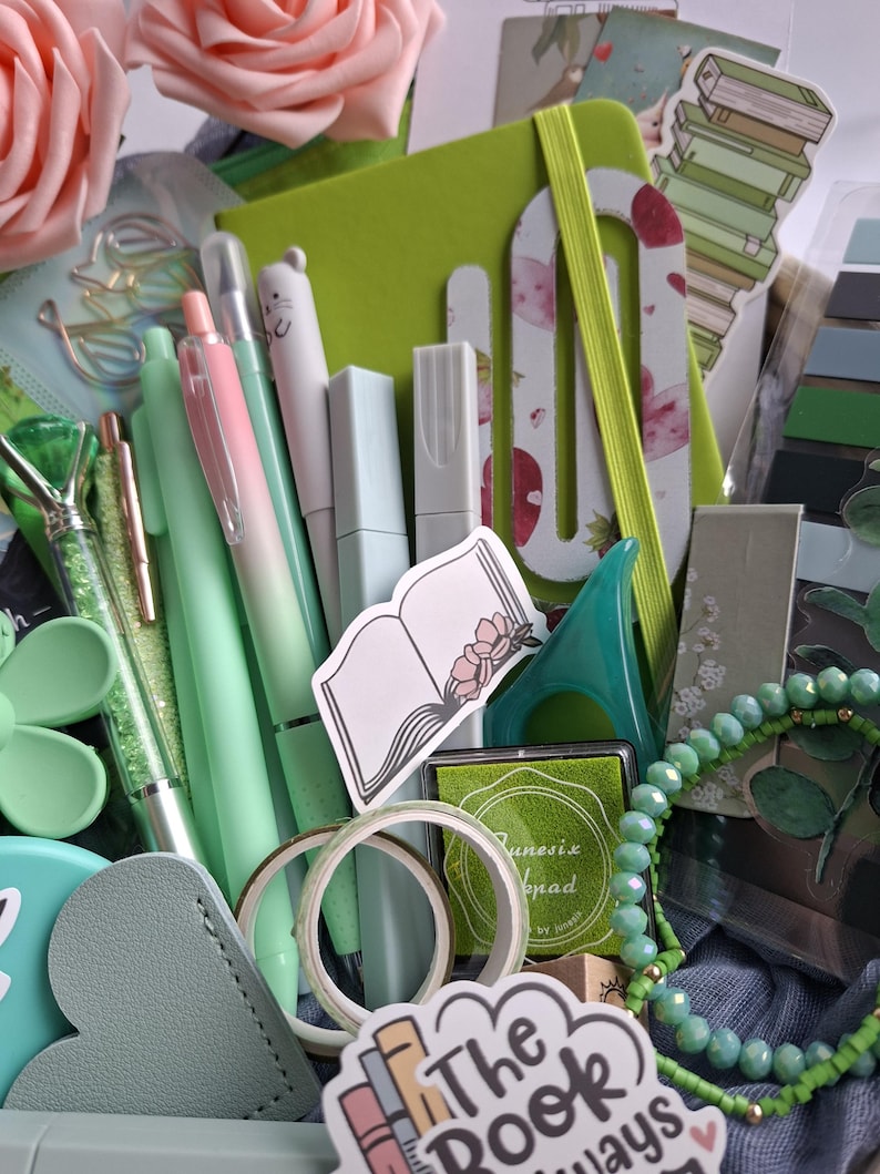 May include: A collection of stationery items in shades of green and pink, including a lime green notebook with an elastic band, various pens, a small ink pad, decorative washi tape, a beaded bracelet, stickers of books, and artificial pink roses.