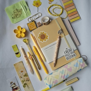 K&ouml;nnte beinhalten: Eine Sammlung von Schreibwaren, darunter ein beigefarbenes liniertes Notizbuch mit Sonnenblumenaufkleber, gelbe Stifte, ein Glitzerstift, ein Federm&auml;ppchen mit Herzmuster, Lesezeichen, Haftnotizen, B&uuml;roklammern, eine gelbe Haarspange und ein Armband.