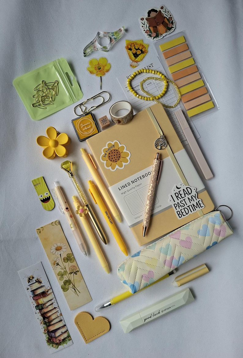 K&ouml;nnte beinhalten: Eine Sammlung von Schreibwaren, darunter ein beigefarbenes liniertes Notizbuch mit Sonnenblumenaufkleber, gelbe Stifte, ein Glitzerstift, ein Federm&auml;ppchen mit Herzmuster, Lesezeichen, Haftnotizen, B&uuml;roklammern, eine gelbe Haarspange und ein Armband.