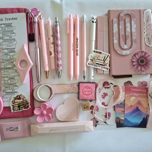 May include: A collection of pink stationery items including a book tracker, pens, a notebook with a large paperclip, sticky notes, a small mirror, a flower-shaped clip, a heart-shaped tray, a stamp pad, a beaded bracelet, and decorative stickers.