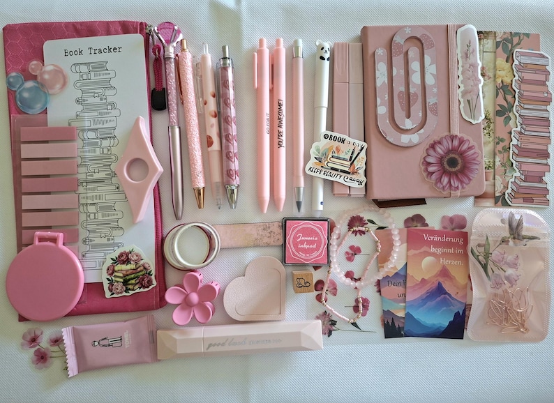May include: A collection of pink stationery items including a book tracker, pens, a notebook with a large paperclip, sticky notes, a small mirror, a flower-shaped clip, a heart-shaped tray, a stamp pad, a beaded bracelet, and decorative stickers.