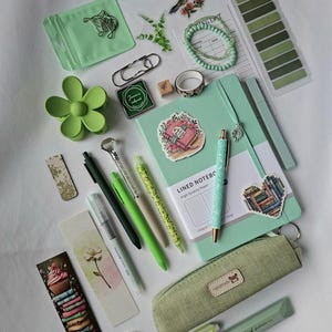 K&ouml;nnte beinhalten: Eine Sammlung von Schreibwaren in Gr&uuml;nt&ouml;nen, darunter ein liniertes Notizbuch, ein Stoffetui mit einem handgefertigten Etikett, verschiedene Stifte, eine blumenf&ouml;rmige Haarspange, Lesezeichen, Aufkleber, ein Perlenarmband und B&uuml;roklammern.