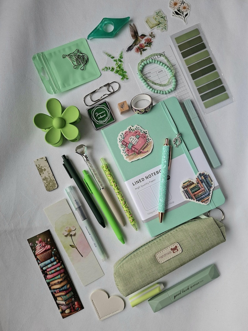 K&ouml;nnte beinhalten: Eine Sammlung von Schreibwaren in Gr&uuml;nt&ouml;nen, darunter ein liniertes Notizbuch, ein Stoffetui mit einem handgefertigten Etikett, verschiedene Stifte, eine blumenf&ouml;rmige Haarspange, Lesezeichen, Aufkleber, ein Perlenarmband und B&uuml;roklammern.