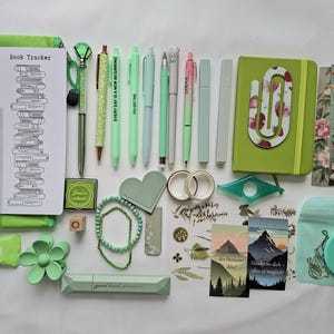 May include: A collection of green stationery items including a book tracker, pens, a notebook with a paperclip, washi tape, stickers, a hair clip, a small mirror, and bookmarks. The items feature various shades of green, floral patterns, and mountain illustrations with German text.