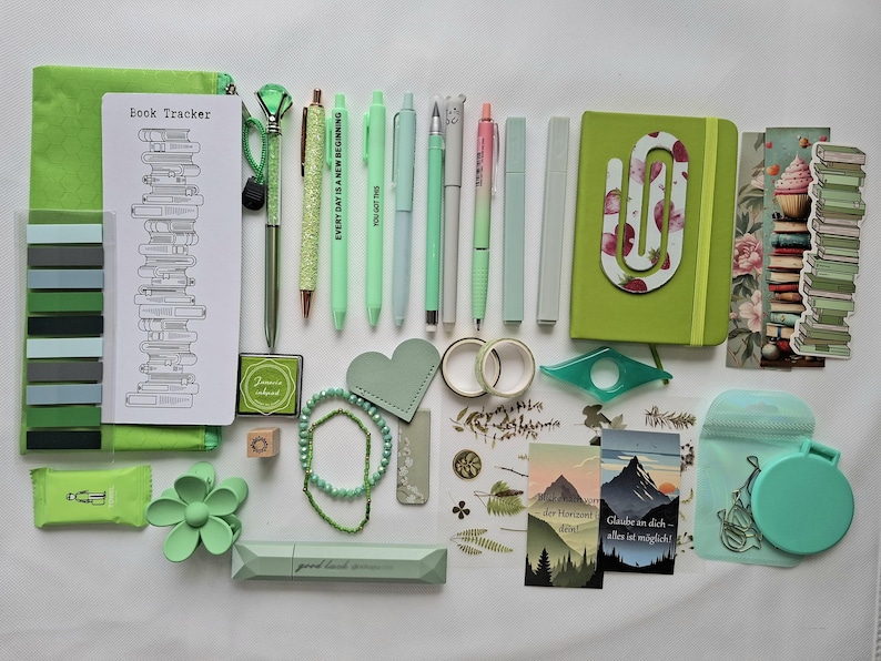 May include: A collection of green stationery items including a book tracker, pens, a notebook with a paperclip, washi tape, stickers, a hair clip, a small mirror, and bookmarks. The items feature various shades of green, floral patterns, and mountain illustrations with German text.