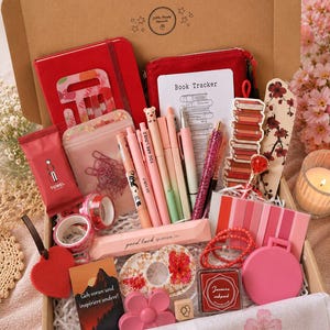 May include: A gift box containing a red notebook, a red pouch, a book tracker card, pens, a stack of books bookmark, a floral bookmark, pink washi tape, a heart ornament, a card with German text, a flower-shaped object, and a pink inkpad.