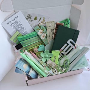 Puede incluir: Una caja de regalo llena de artículos de papelería verdes y en tonos pastel. Incluye bolígrafos, un cuaderno, marcadores, un clip con forma de flor y una pequeña bolsa verde. La caja también contiene una tarjeta que dice "Book Tracker".