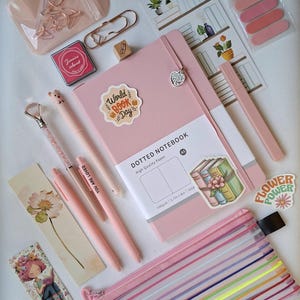 K&ouml;nnte beinhalten: Ein rosafarbenes A5-Notizbuch mit Punkten und Herzanh&auml;nger, dazu rosa Stifte, ein Stift mit Kristallspitze, ein Netz-Etui mit Regenbogenstreifen, Lesezeichen, Aufkleber mit "I LOVE READING" und "World BOOK Day" sowie ros&eacute;goldene B&uuml;roklammern.