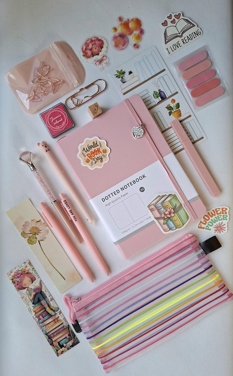 K&ouml;nnte beinhalten: Ein rosafarbenes A5-Notizbuch mit Punkten und Herzanh&auml;nger, dazu rosa Stifte, ein Stift mit Kristallspitze, ein Netz-Etui mit Regenbogenstreifen, Lesezeichen, Aufkleber mit "I LOVE READING" und "World BOOK Day" sowie ros&eacute;goldene B&uuml;roklammern.