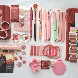 May include: A collection of stationery items in shades of pink and red, including a notebook, pens, washi tape, stickers, a book tracker, a pencil case, sticky notes, a flower-shaped hair clip, a small mirror, and a beaded bracelet.