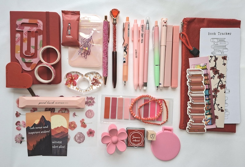 May include: A collection of stationery items in shades of pink and red, including a notebook, pens, washi tape, stickers, a book tracker, a pencil case, sticky notes, a flower-shaped hair clip, a small mirror, and a beaded bracelet.