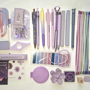 May include: A collection of lavender-themed stationery including a dotted notebook, washi tape, pens, a pencil case, sticky notes, a beaded bracelet, a flower-shaped hair clip, stickers, and a book tracker with a stack of books illustration.