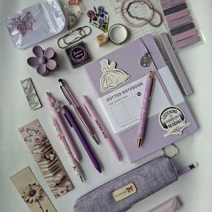 K&ouml;nnte beinhalten: Eine Sammlung von Schreibwaren in Lila- und Lavendelt&ouml;nen, darunter ein punktiertes Notizbuch, ein Stiftem&auml;ppchen aus Stoff, mehrere Stifte, Lesezeichen, Aufkleber, eine blumenf&ouml;rmige Haarspange, B&uuml;roklammern und eine Leseliste.