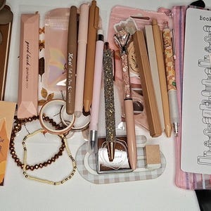 May include: A collection of stationery items including a brown notebook with a flower clip, various pens in shades of beige and pink, a glittery pen, a beaded bracelet, a book tracker card, and a clear pencil case with a stack of books illustration.