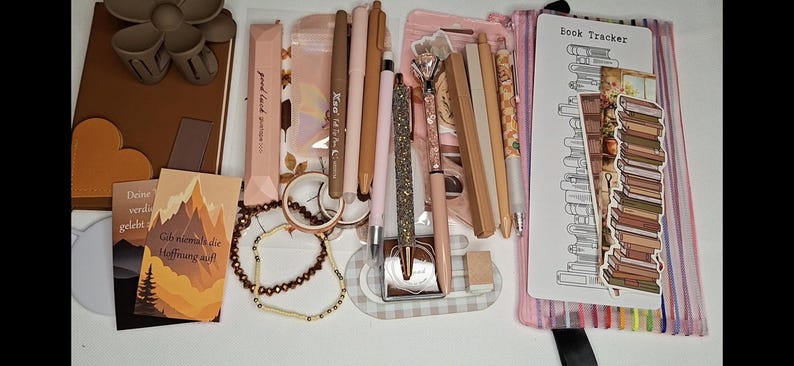 May include: A collection of stationery items including a brown notebook with a flower clip, various pens in shades of beige and pink, a glittery pen, a beaded bracelet, a book tracker card, and a clear pencil case with a stack of books illustration.