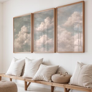 Könnte beinhalten: Drei gerahmte Kunstdrucke mit Wolkenlandschaft und hellen Holzrahmen hängen über einer Holzbank mit weißen Kissen. Das Kunstwerk zeigt flauschige weiße und rosa Wolken vor einem blau-grauen Himmel. Ein geflochtener Korb und ein gestreiftes Kissen befinden sich auf der Bank.